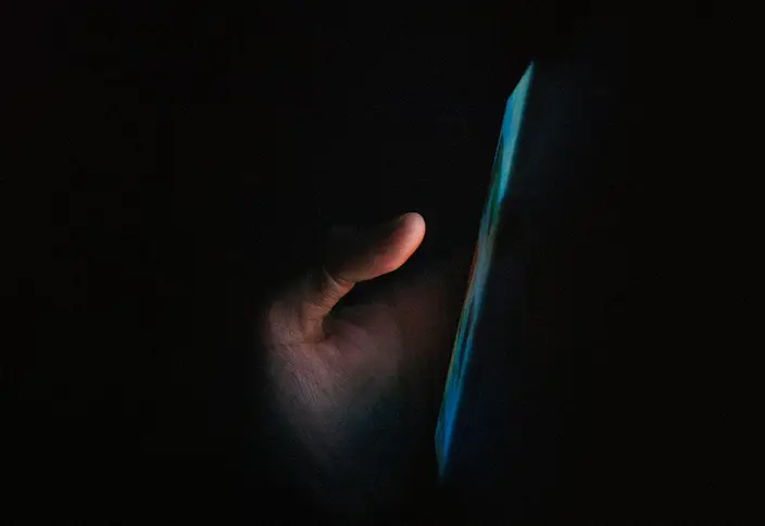 Mano acercándose a la pantalla azul encendida de un teléfono en una habitación a oscuras